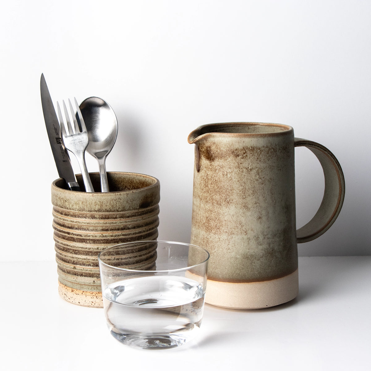 Een keramische beker met bestek staat naast de AB Ceramics Karaf Wit Mos 70 cl en een helder glas water, allemaal gerangschikt op een wit oppervlak tegen een witte achtergrond.