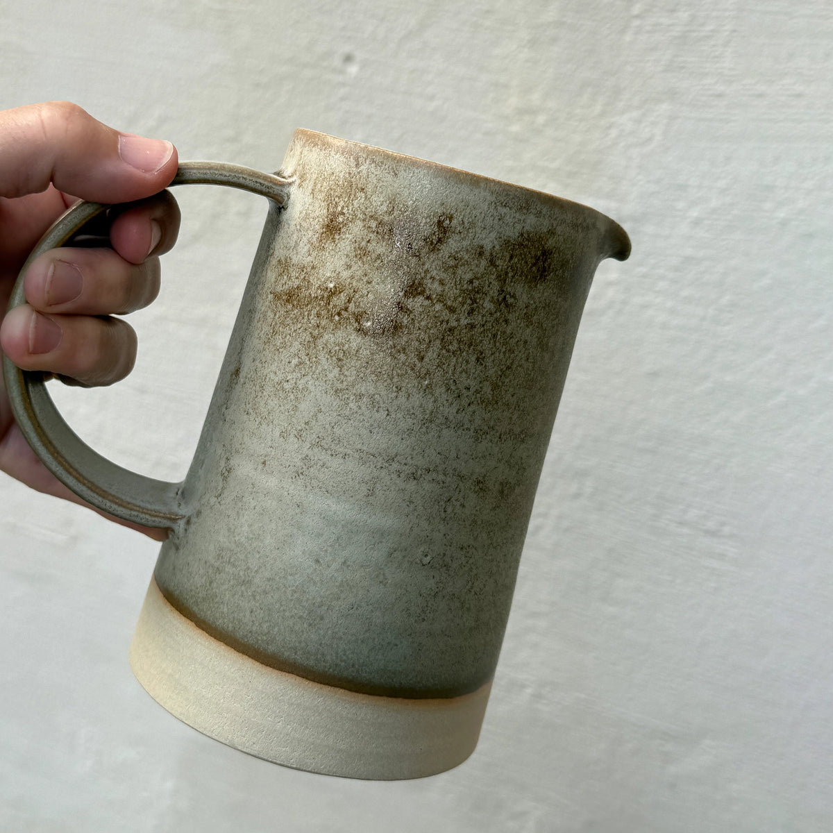 Een hand houdt de AB Ceramics Karaf Wit Mos 70 cl vast, gemaakt van witte klei met een verloop van aardetinten - beige aan de basis, bruin aan de bovenkant en subtiel moskleurig glazuur - tegen een effen lichte achtergrond.