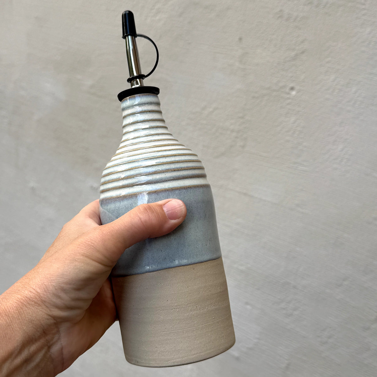 Een hand houdt de AB Ceramics Olijfolie Fles Ribbel Wit Opaal 45 cl vast, met een geribbelde witte opalen bovenkant, gladde onderste helft en een metalen schenktuit, tegen een lichtgrijze wand met textuur.
