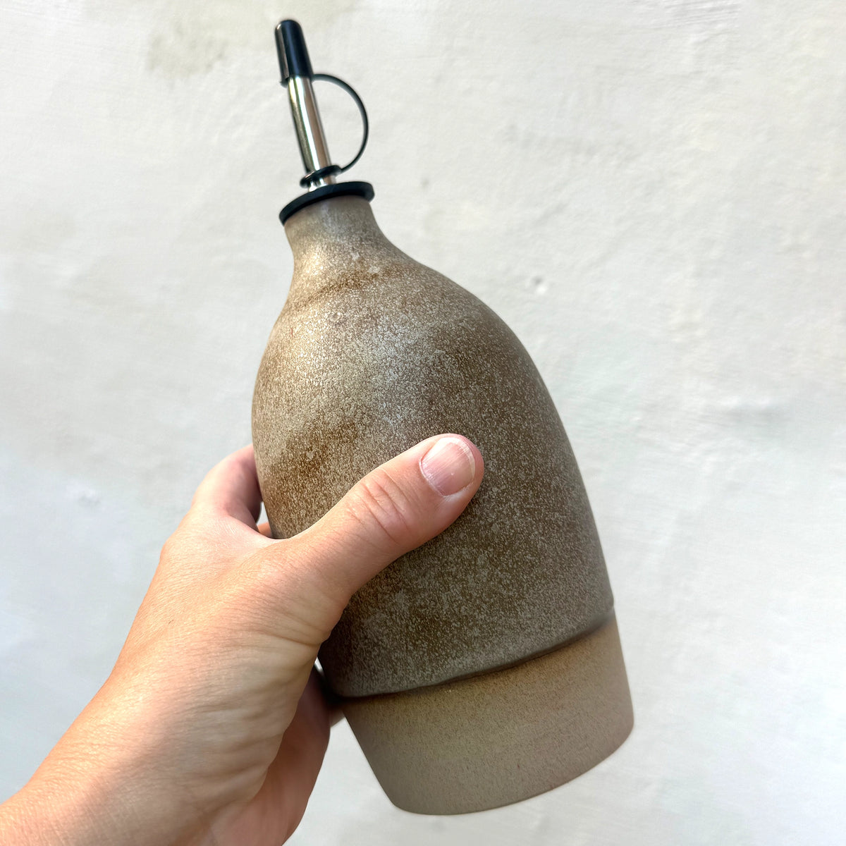 Een hand met de AB Ceramics Olijfolie Fles 2 Toffee Mos 65 cl met een metalen schenktuit, perfect voor olie of azijn, tegen een lichte achtergrond-een stijlvolle aanvulling op je keuken decoratie van AB Ceramics.