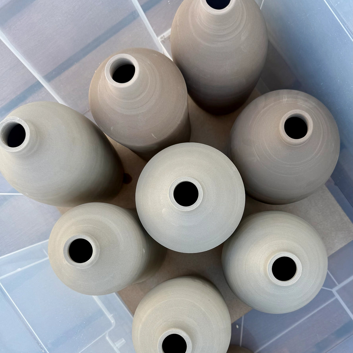 Bovenaanzicht van negen handgedraaide keramiekvazen met smalle openingen, gerangschikt in een transparante container. De ongeglazuurde, zachte neutrale tinten doen denken aan de AB Ceramics Olijfolie Fles Toffee Opaalgroen 55 cl.