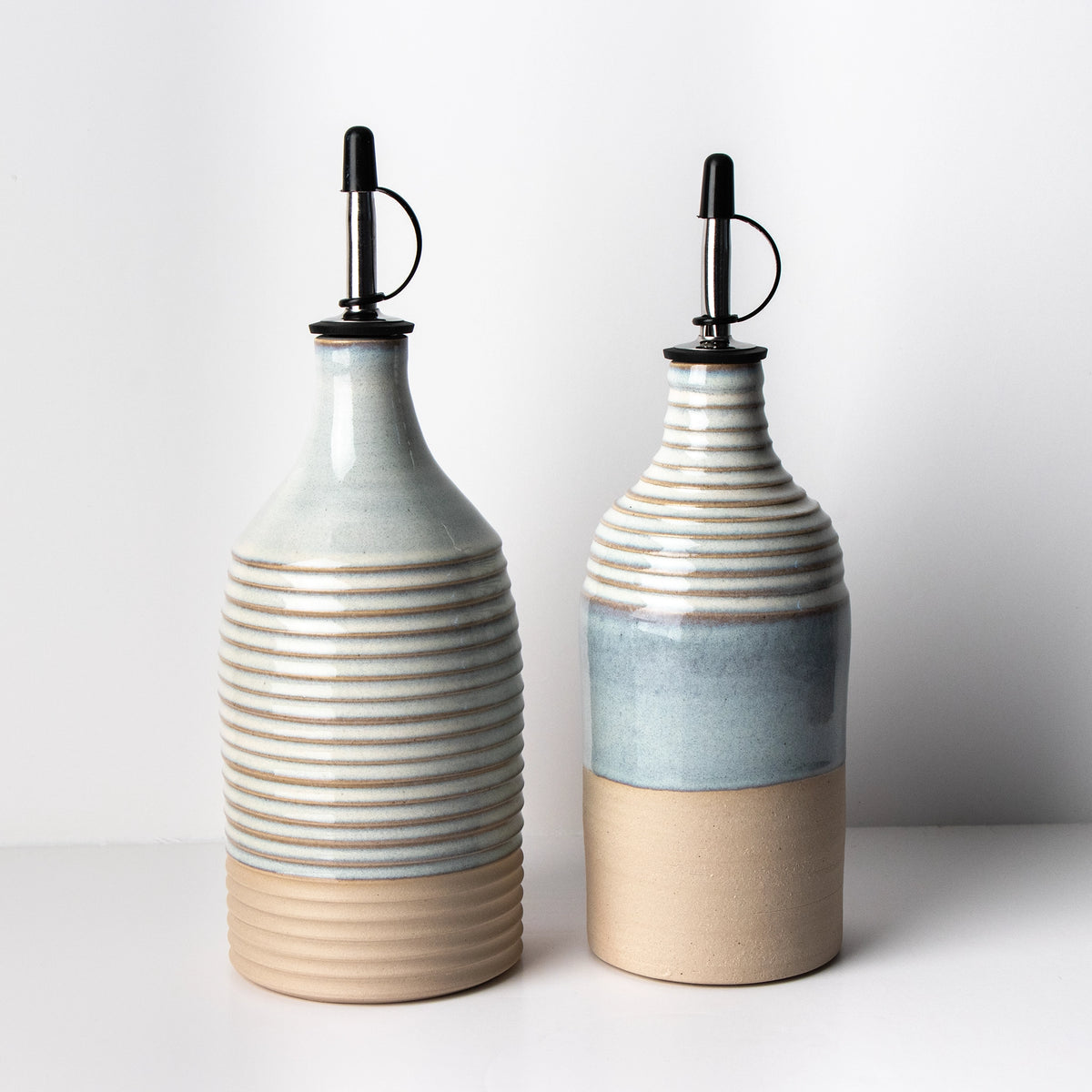 Twee AB Ceramics Olijfolie Fles Ribbel Wit Opaal 65 cl dispensers met geribbelde textuur, neutrale tinten en zwarte schenktuiten staan naast elkaar en benadrukken hun moderne minimalistische ontwerp tegen een effen witte achtergrond.