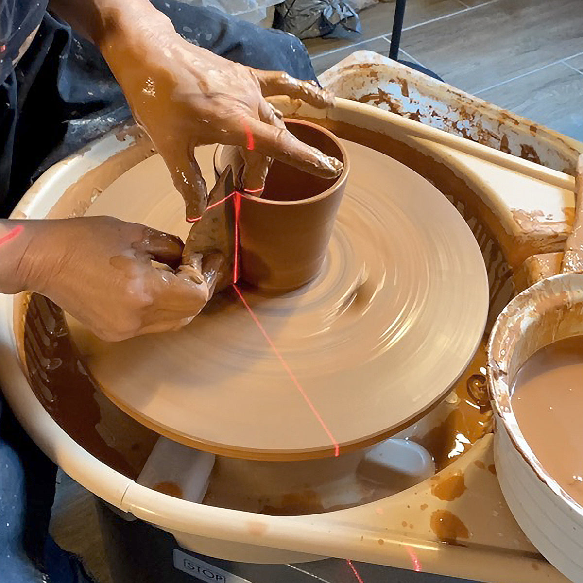 Een ambachtsman vormt de AB Ceramics Beker Wit Mat Wit 20 cl op een draaiende pottenbakkersschijf. Witte steengoed klei wordt met de hand geleid terwijl een rode laserlijn voor uitlijning zorgt. Gereedschap van AB Ceramics is zichtbaar rond het wiel.