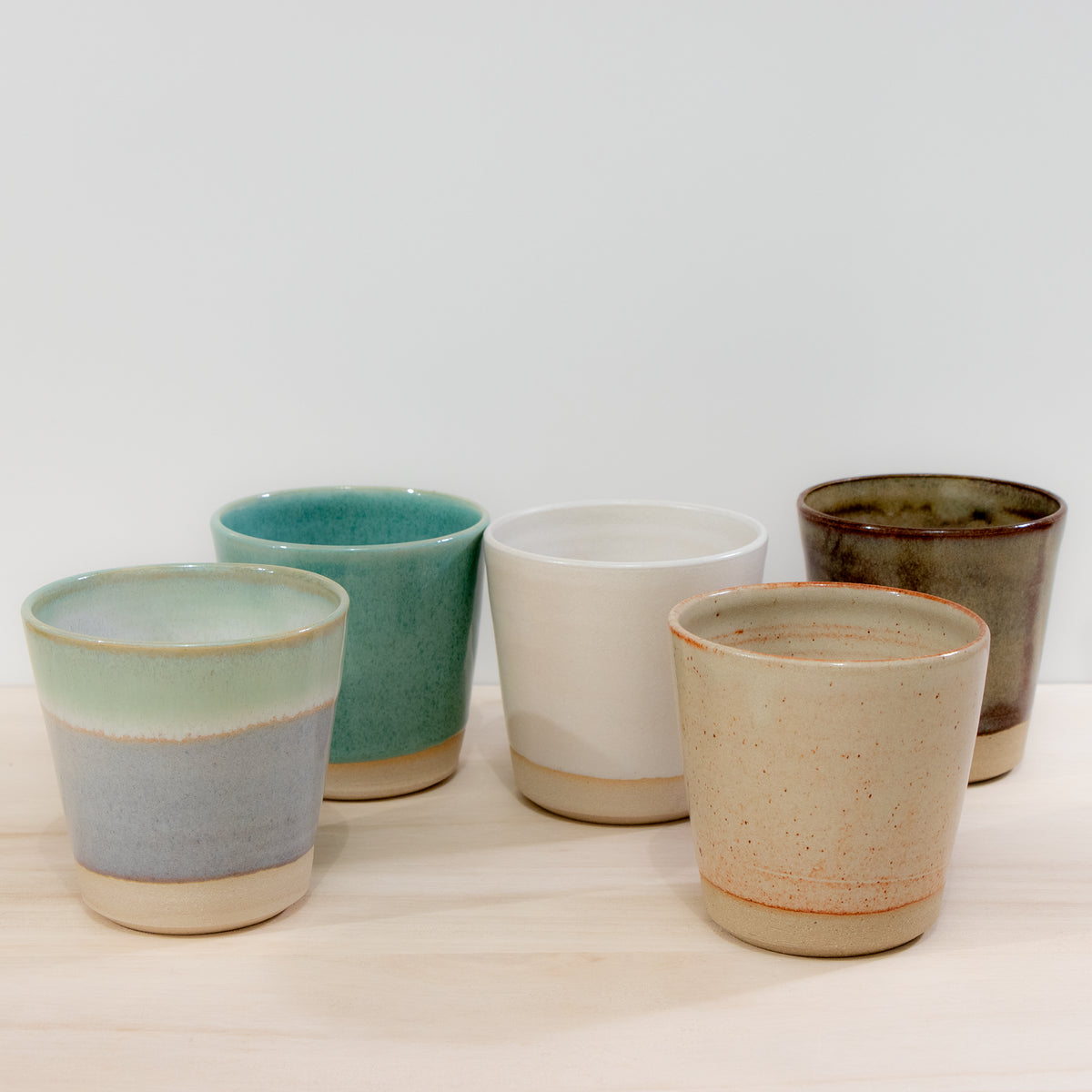 De AB Ceramics Beker Wit Opaal/Lichtgroen 20 cl, een uniek handgemaakte beker van wit steengoed, staat op een licht houten oppervlak tegen een effen witte achtergrond.