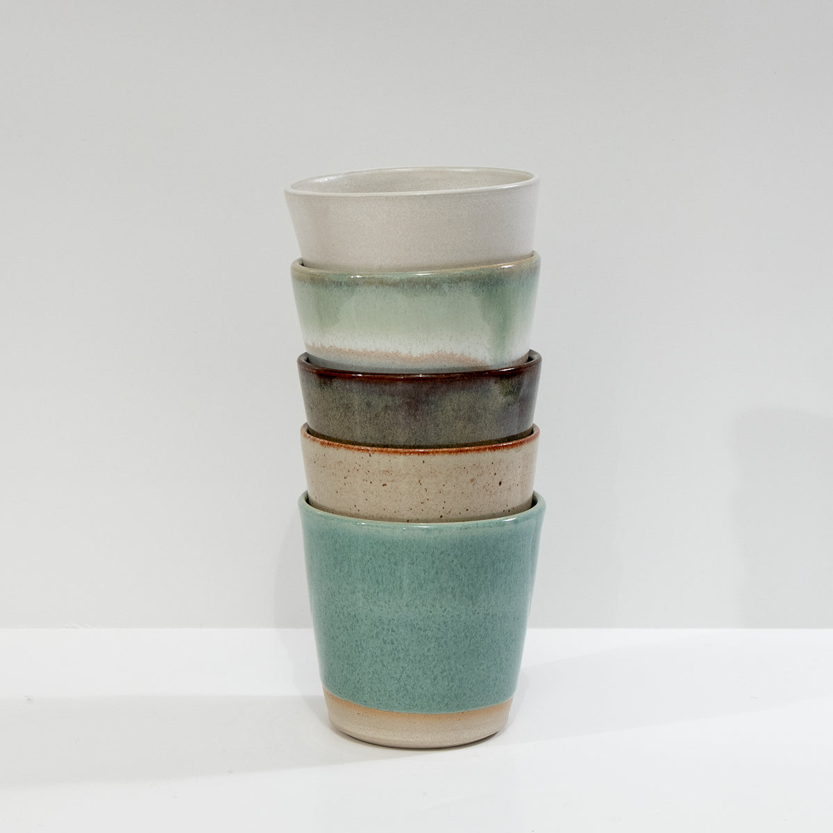 Vier AB Ceramics Beker Wit Opaal/Lichtgroen 20 cl kopjes, elk met een unieke handgeglazuurde witte of lichtgroene afwerking op wit steengoed klei, zijn gestapeld in een minimalistische stijl tegen een effen witte achtergrond.