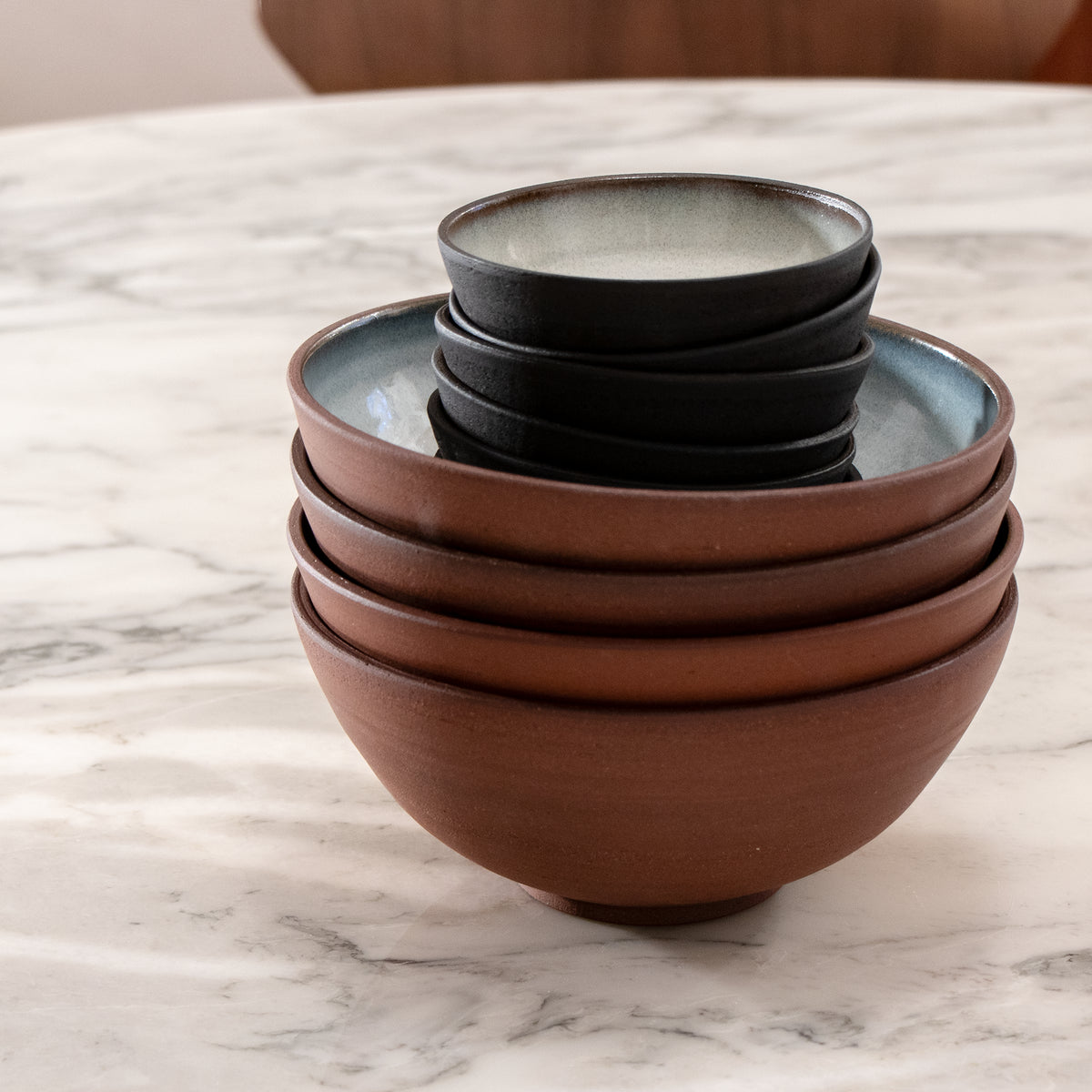 Een stapel van vier Kom Rood Opaal D18 kommen van AB Ceramics, elk met een kleinere donkere noedelkom erin genesteld, geplaatst op een wit marmeren tafelblad.