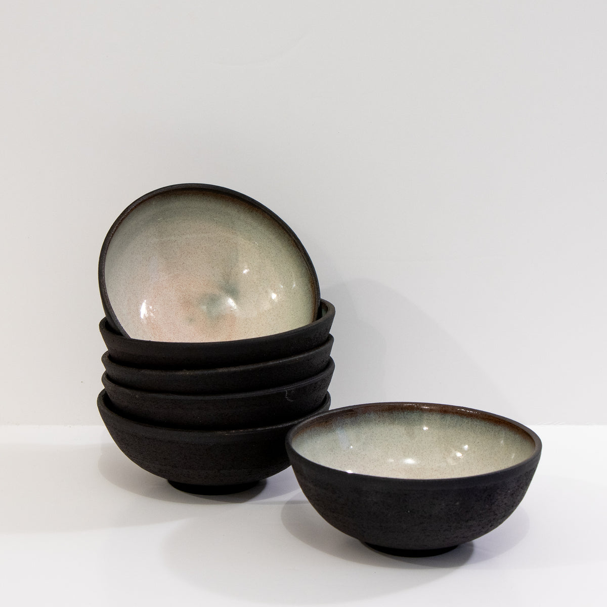 Links staan vijf AB Ceramics Mini Kom Zwart Opaal D10,5 kommen met een glanzende binnenkant, rechts één op zichzelf staande kom tegen een effen witte achtergrond.