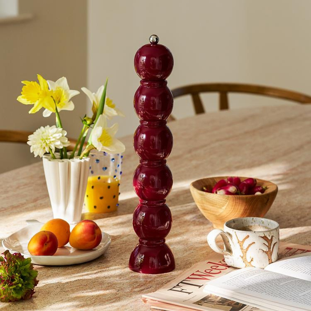 Een hoge, glanzende Addison Ross Bobbin Zout-en Pepermolen Kersenrood 35 cm staat op een zonovergoten tafel met narcissen in een vaas, een houten schaal met kersen en abrikozen, een stippenglas, een mok en een open tijdschrift.