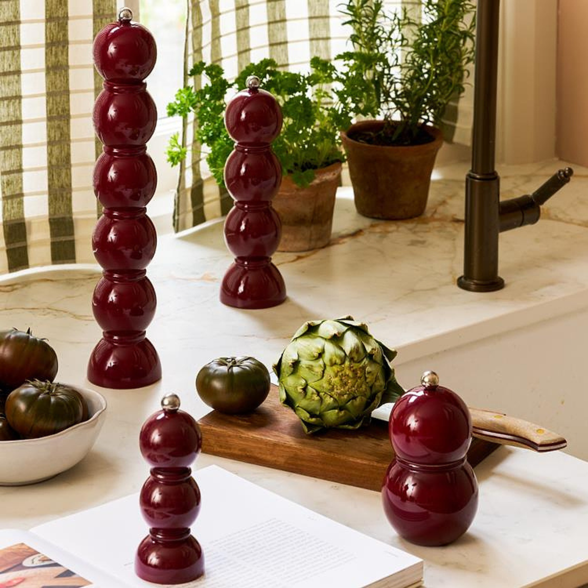 Een 35 cm kersenrood Addison Ross Bobbin zout-en pepermolen staat op een aanrecht met heirloomtomaten, artisjok, kruiden in potten, een open boek en een gestreept gordijn op de achtergrond.