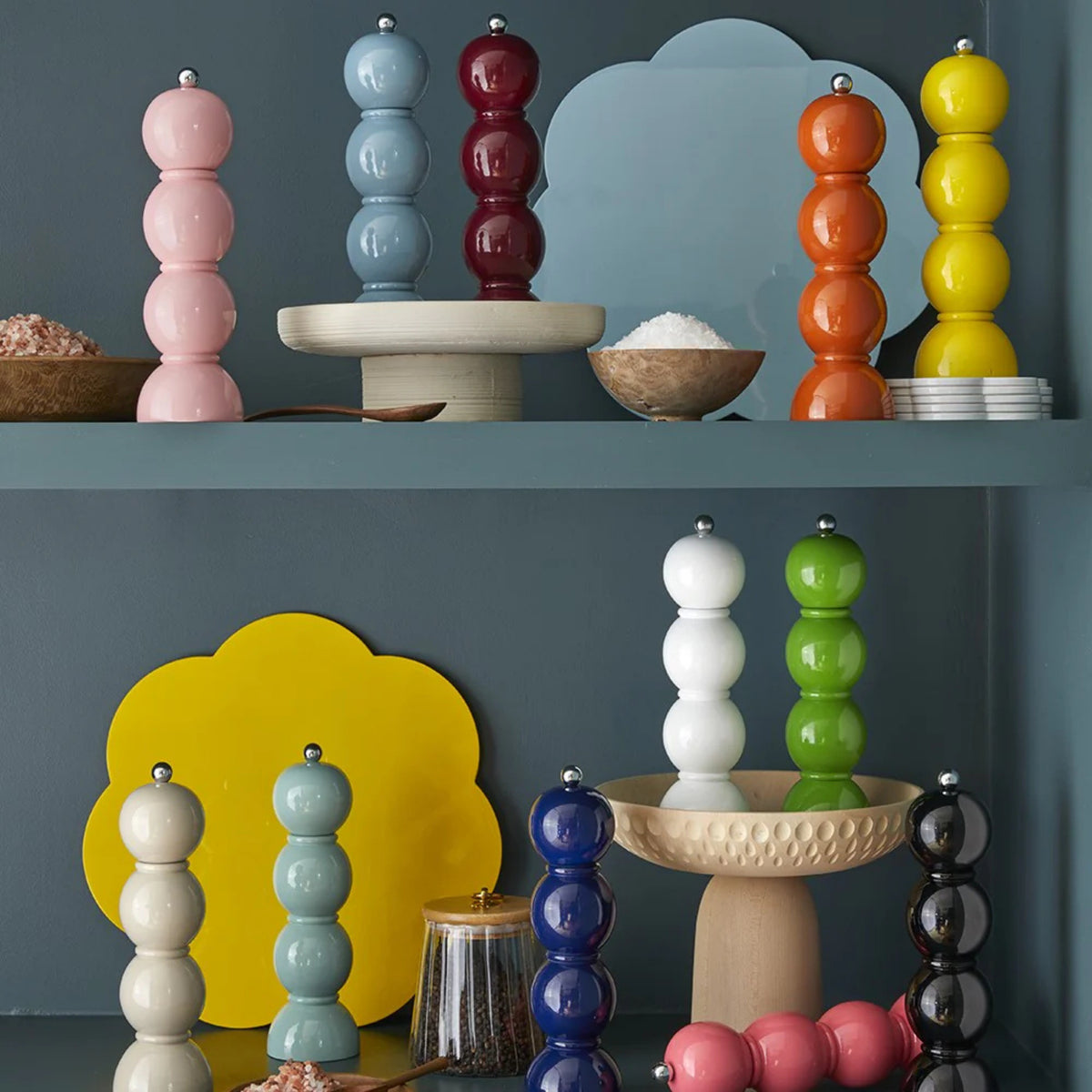 De Addison Ross Bobbin Zout-en Pepermolen Racing Groen 24 cm staat mooi op blauwe planken, omringd door zoutkommen, kleine bordjes en gele en lichtblauwe wolkvormige borden voor een levendige, moderne keukenlook.
