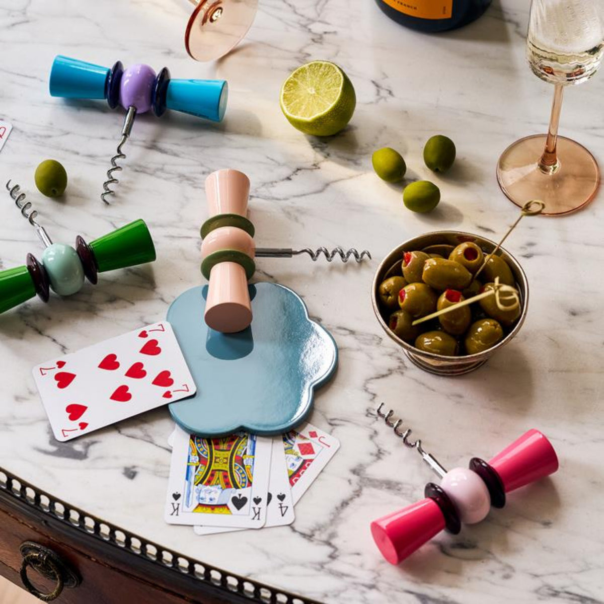 Een marmeren tafel met de Addison Ross Bowtie Kurkentrekker Leaf & Eau de Nil, speelse groene kurkentrekkers, groene olijven, speelkaarten, een gehalveerde limoen en een glas wijn voor een casual en toch elegante vibe.