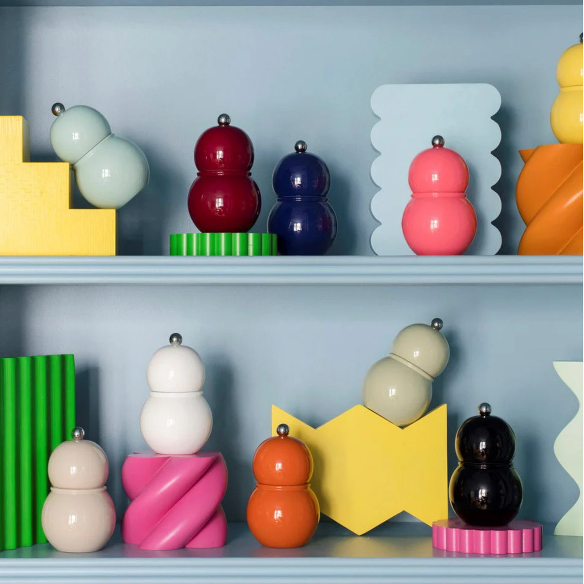 Op een plank staan de Addison Ross Chubby Zout-en Pepermolen Oranje 12 cm en andere molens, geaccentueerd door gele, groene en roze geometrische vormen op een blauwe achtergrond - ideaal voor elke Addison Ross design keukengerei fan.
