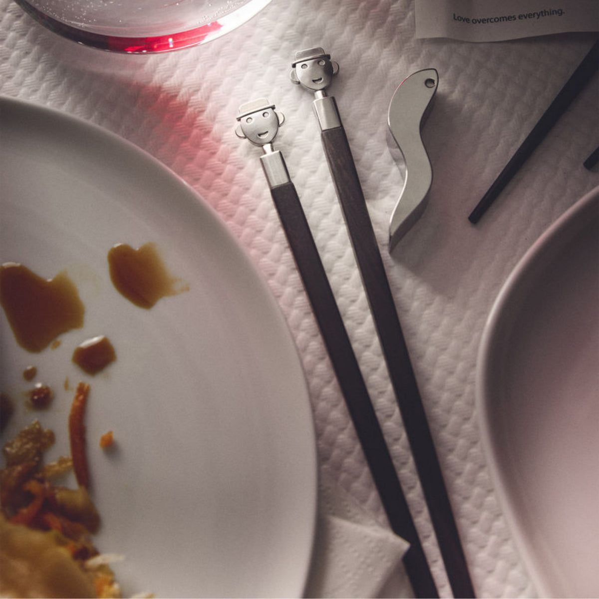 Twee Alessi Alessandro eetstokjes + Houder van Alessi, met cartoonachtige gezichten geïnspireerd door Mendini, rusten op een wit oppervlak met textuur naast tafelaccessoires: een zilveren flesopener, een bord met eten en saus, een rood drankje in een glas en een zichtbare kaart.