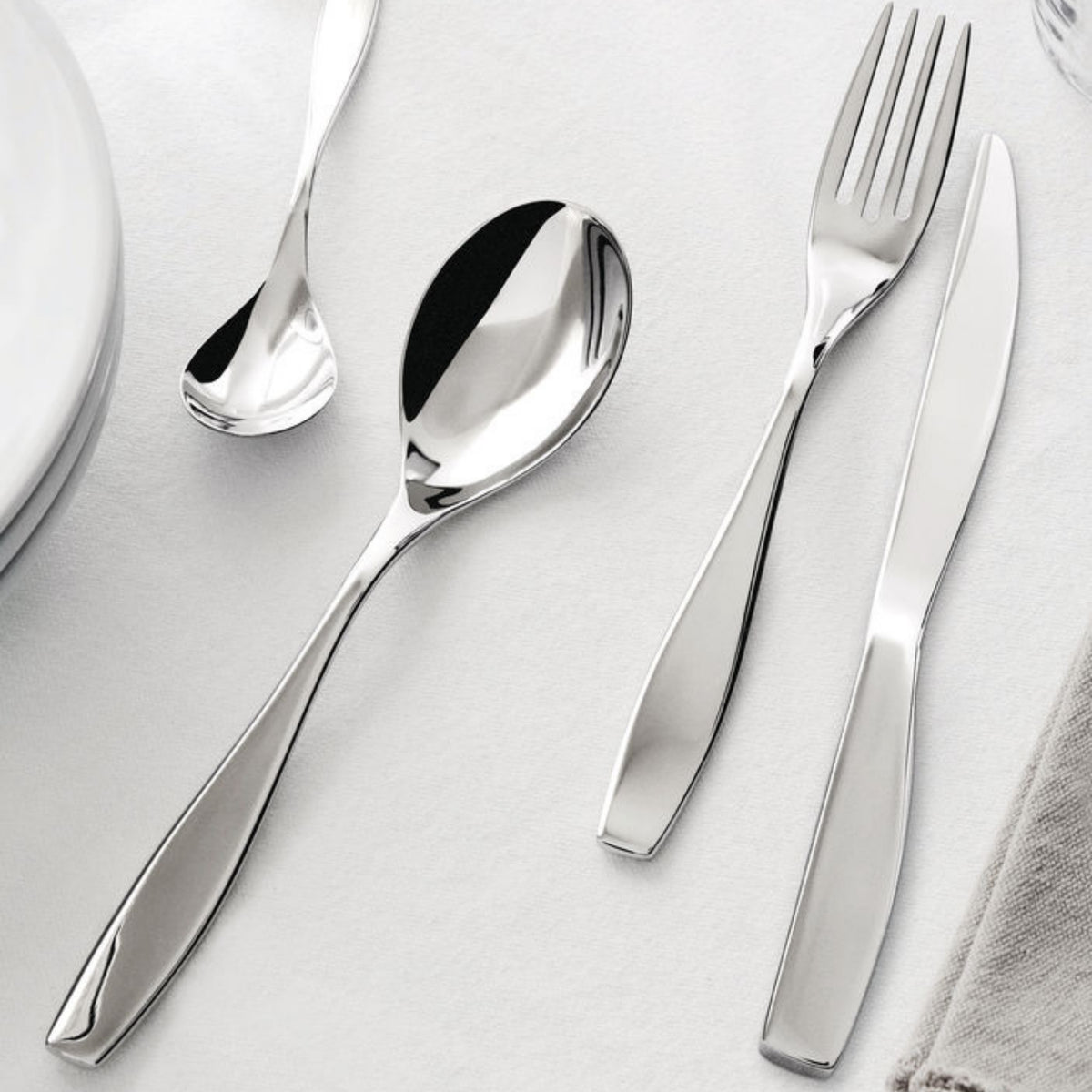 Een Alessi Duna Bestek 24-delig servies met Italiaans design staat op een wit tafelkleed naast een bord en servet.