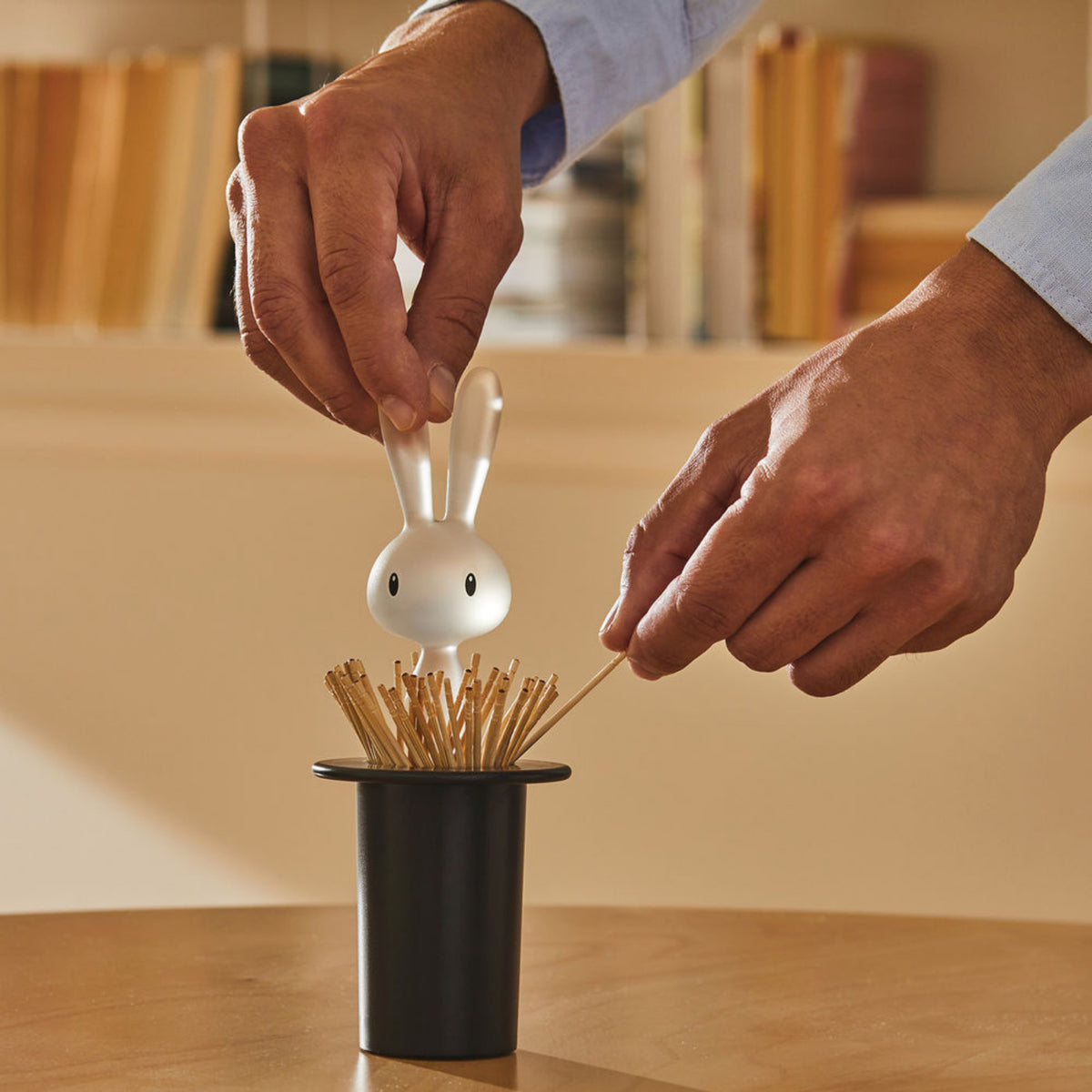 Een persoon pakt een tandenstoker uit de Alessi Magic Bunny Tandenstokerhouder Zwart, met een wit konijnvormig blad van Alessi, geplaatst op een tafel met vage planken en boeken op de achtergrond.