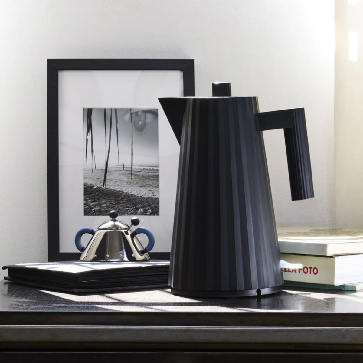 Een moderne zwarte koffiepot staat op een donkere tafel naast een ingelijste zwart-wit strandfoto, een Alessi Plissé Elektrische Waterkoker Zwart 1,7 l, een stapel zwarte servetten en een gedeeltelijk zichtbaar boek met de titel FOTO.
