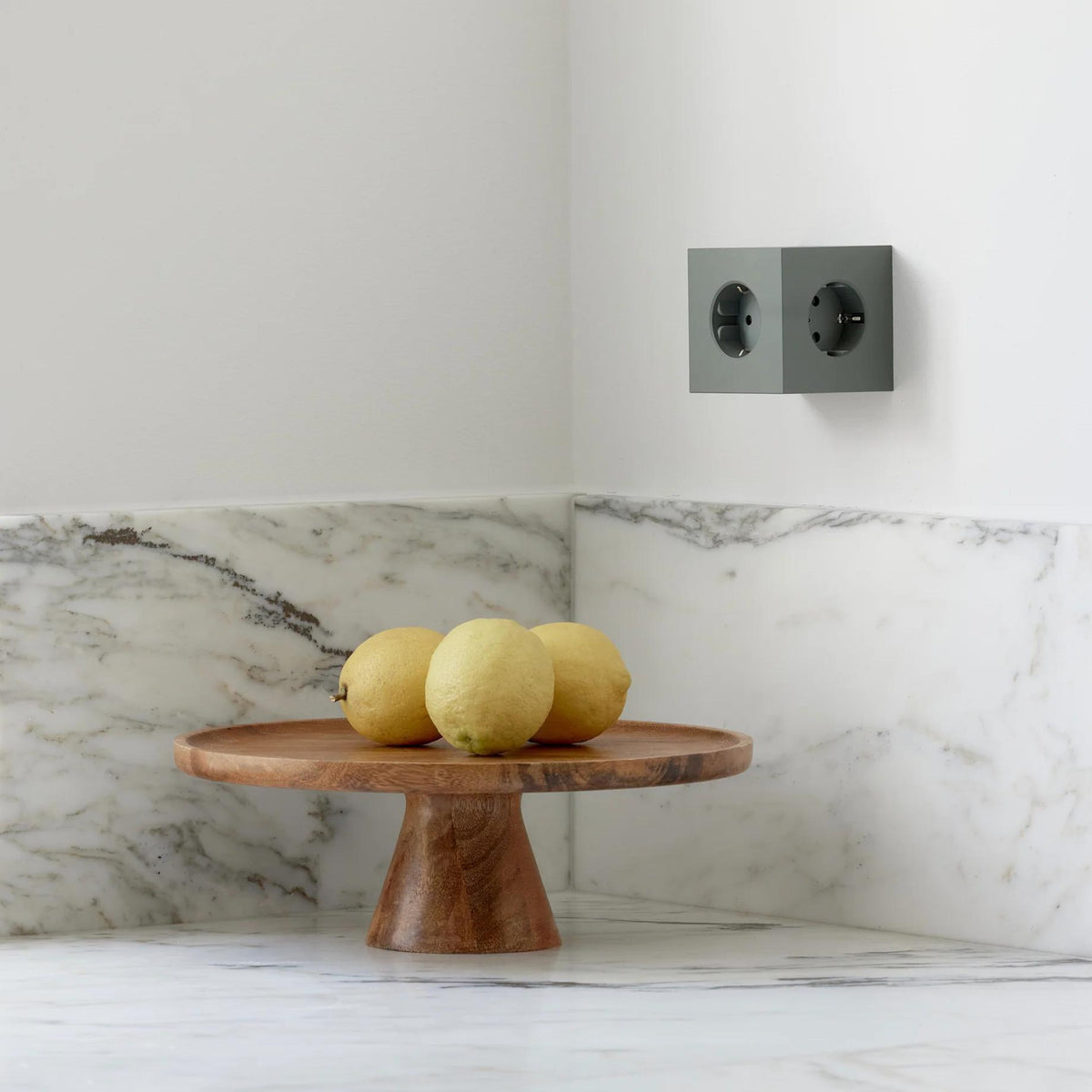 Een houten voetstuk met drie citroenen staat op een wit marmeren aanrecht. Daarboven is een Avolt Square 2 USB-C Stekkerdoos Eik Groen van Avolt gemonteerd naast het dubbele stopcontact.
