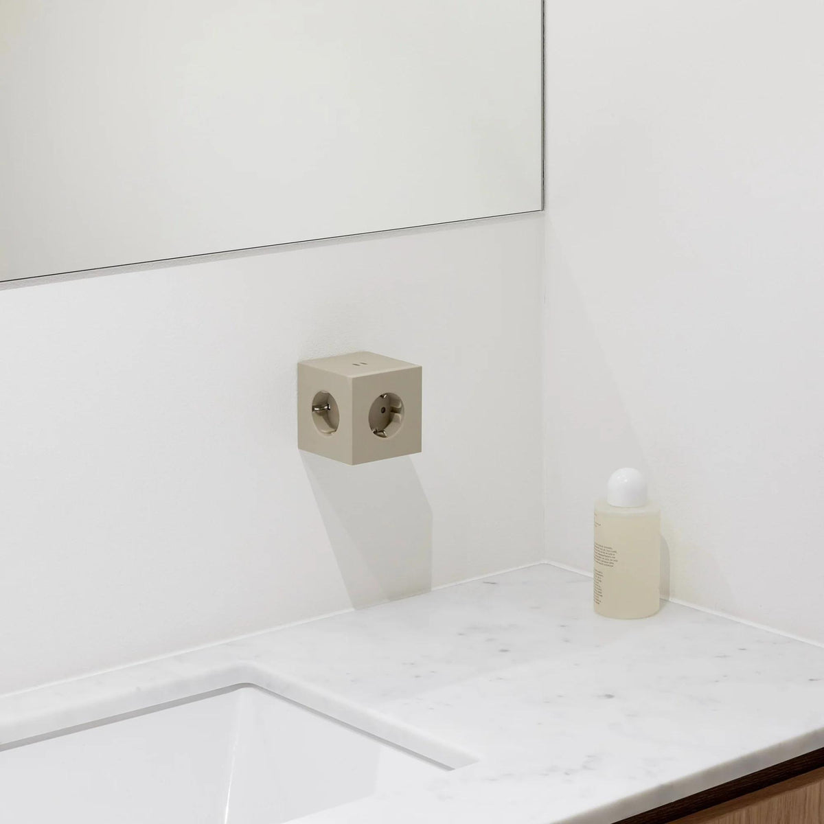 Een minimale badkamer heeft een wit marmeren aanrechtblad met een vierkante wastafel, een beige fles vloeibare zeep en de Avolt Square 2 USB-C Stekkerdoos Beige van Avolt aan de muur onder een grote spiegel.