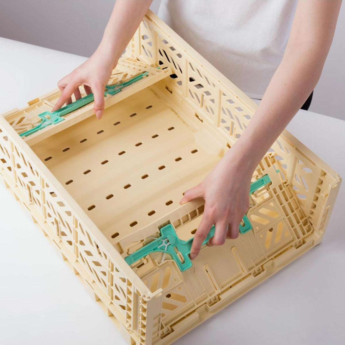 De handen van een persoon vouwen langs de zijkanten van een beige Aykasa Vouwkrat Maxi Comfort Lock Banana / Munt opbergkist van Aykasa, met groene scharnieren, geplaatst op een wit oppervlak.