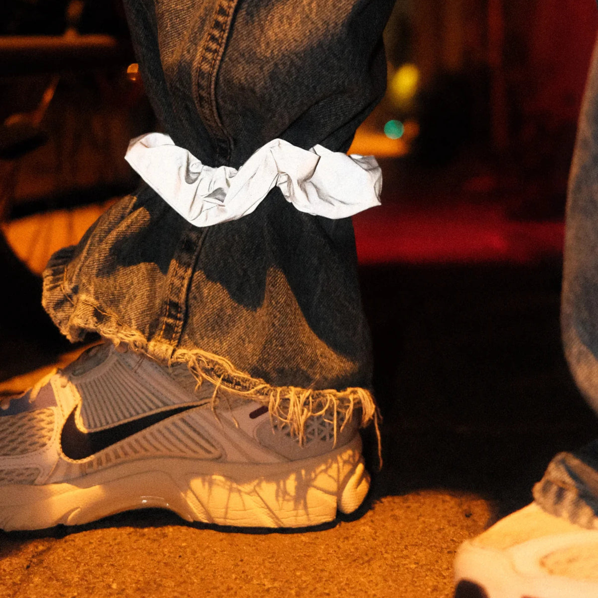 Een persoon in een gerafelde blauwe spijkerbroek en witte Nike sneakers draagt een Caro Antwerp Caro The Scrunch Club Scrunchie 2 st. om één broekspijp. De schemerige scène heeft een warme, stemmige sfeer.