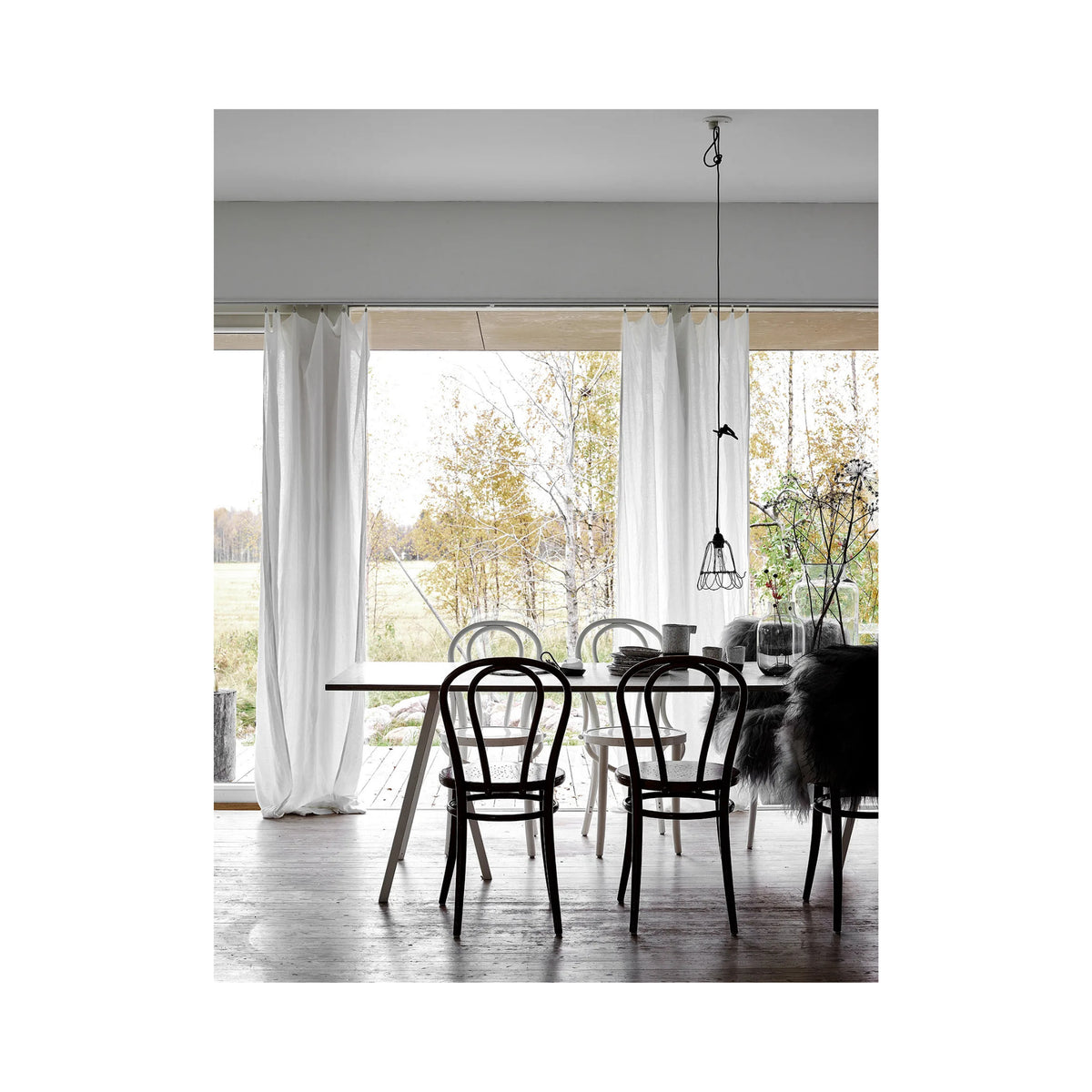 Een minimalistische eetkamer met een witte tafel, zwarte stoelen en grote ramen is gevuld met licht - perfect om het Cozy Publishing Happy Homes Creative Boek door te bladeren in een uitnodigende Scandinavisch geïnspireerde ruimte.