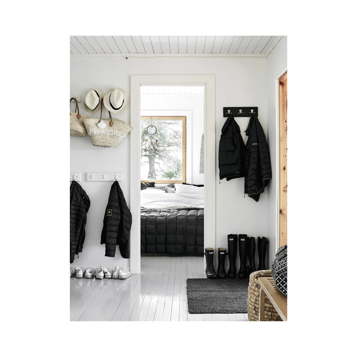 Een lichte entree met witte muren en vloeren, zwarte laarzen en jassen op rekken, hoeden op wasknijpers - een scène rechtstreeks uit het Happy Homes Hideaways Boek van Cozy Publishing - leidt naar een gezellige slaapkamer met een groot raam en een bed met daarop een donker dekbed.