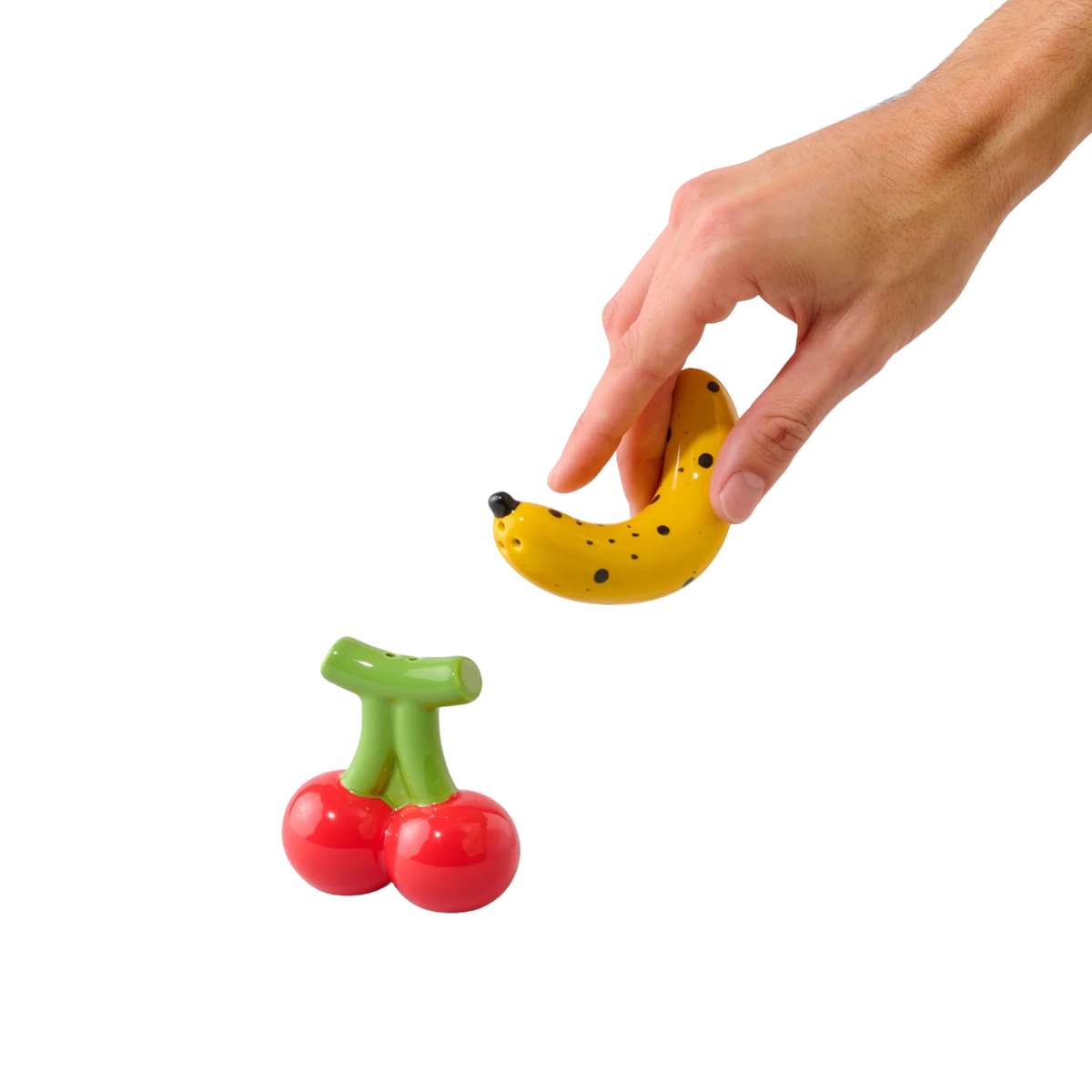Een hand houdt een kleine gele Fluid Market Zout- en Pepervaatjes Banaan vast boven rode Kers op een witte achtergrond, die lijkt op een speels peper- en zoutstel van Fluid Market.