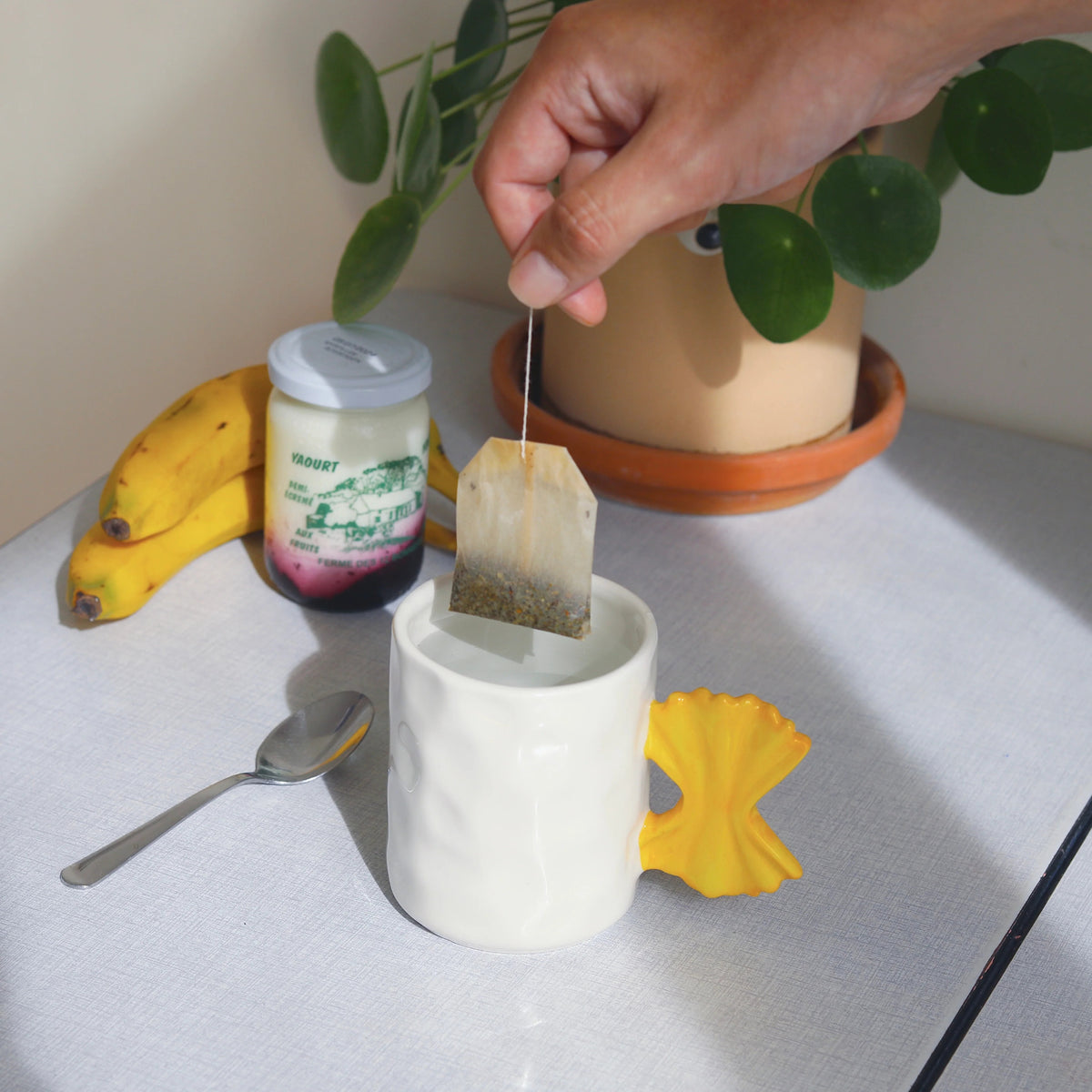 Een hand doopt een theezakje in de Fluid Market Mok Pasta, een witte mok van keramiek met een geel strikhandvat. Ernaast liggen een lepel, yoghurt, twee bananen en een plant in pot - een origineel cadeau voor theeliefhebbers thuis of bij Fluid Market.