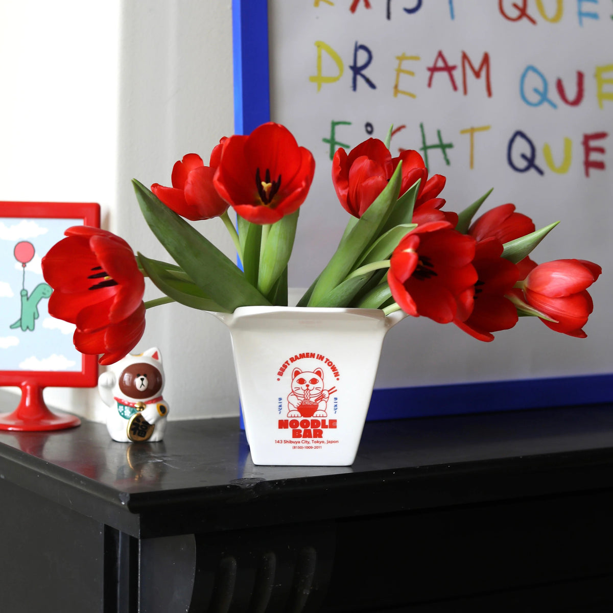 Een Fluid Market Ramen Pot in wit van Fluid Market houdt rode tulpen op een zwarte schouw, naast een maneki-neko kattenbeeldje en ingelijste levendige kunst met speelse tekst-een levendige display voor fans van design en keramieken objecten.