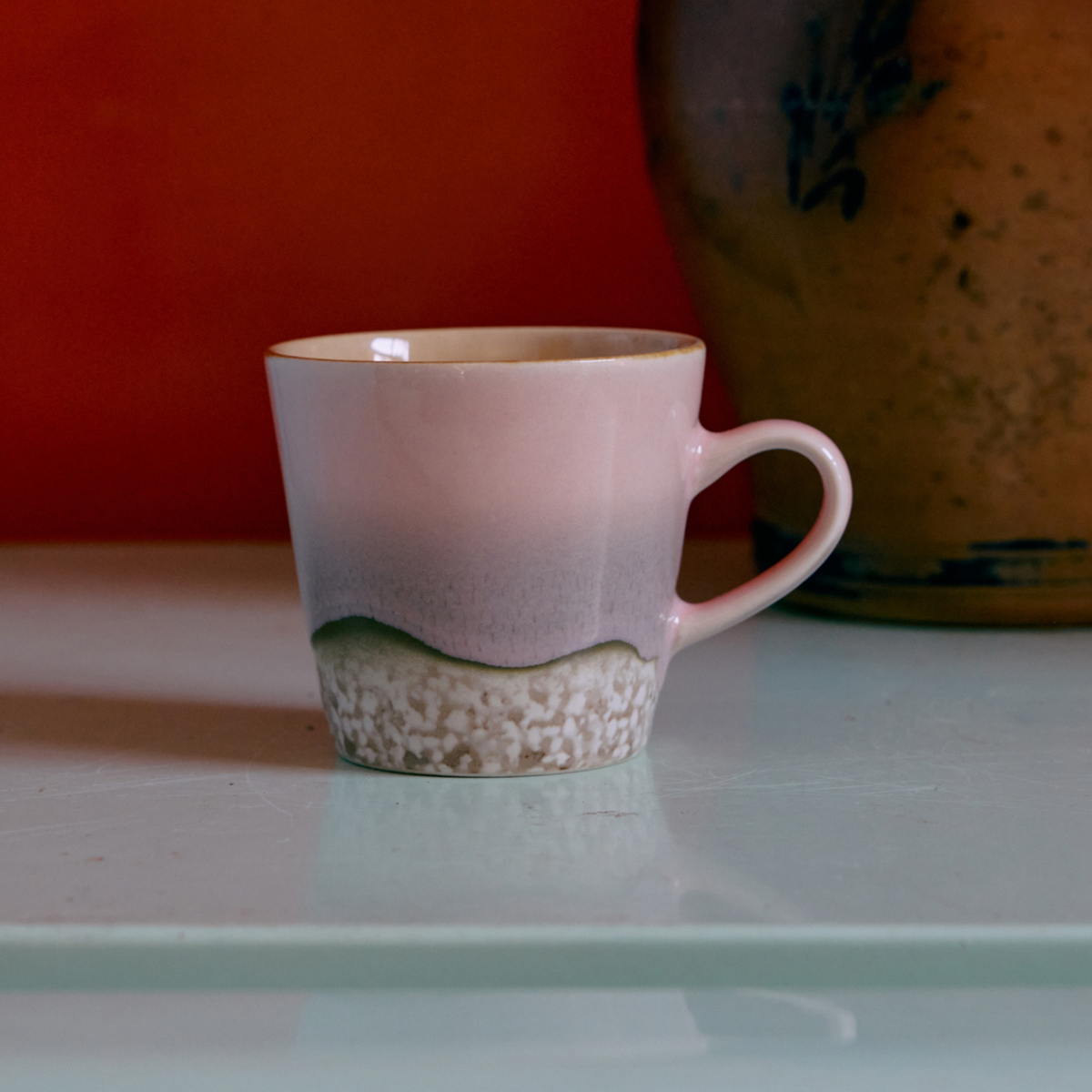 De HK Living 70s Ceramics Americano Mok Muse heeft een zacht lichtroze tot beige kleurverloop met een gestructureerde witte en groenachtige basis. De mok wordt afgebeeld op een lichtgekleurde ondergrond, met op de achtergrond een bruine aardewerken vaas die gedeeltelijk zichtbaar is.
