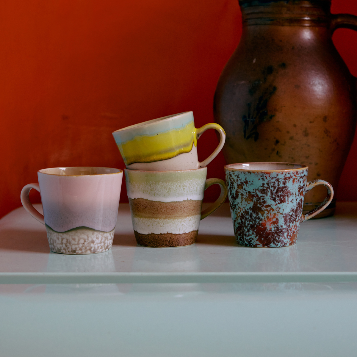 Vier HK Living 70s Ceramics Americano Mok Umber-mokken, met levendige kleuren, staan opgesteld op een licht oppervlak voor een grote bruine kan, tegen een oranje muur. Eén mok is op een andere gestapeld, wat zorgt voor een speelse HK Living-presentatie.