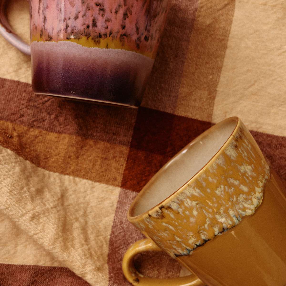 De HK Living 70s Ceramics Americano Mok Sunbeam is voorzien van gevlekte, aardse glazuren in roze, paars, geel en bruin, afgezet tegen een bruin-beige geruite stof — wat de charme van een retro koffiemok oproept.