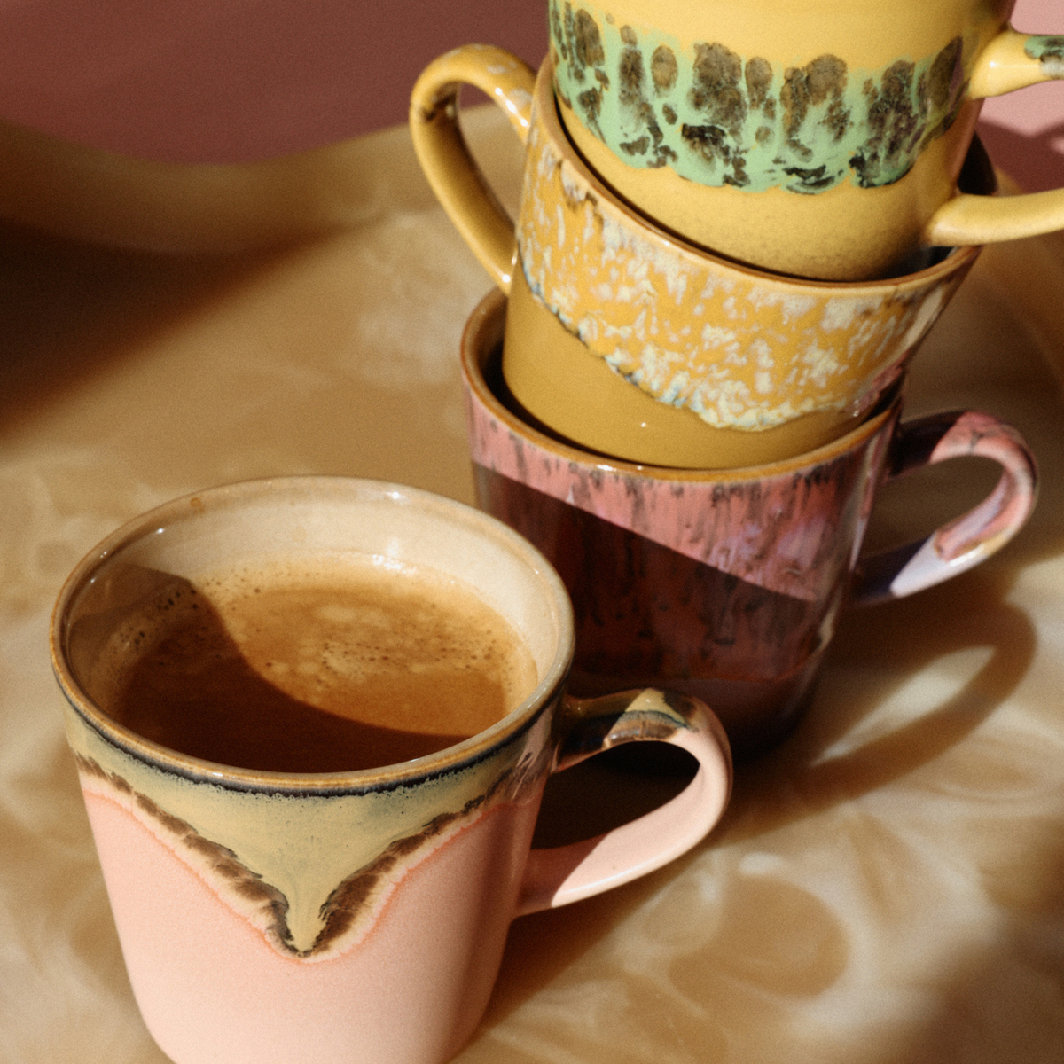 Vier HK Living 70s Ceramics Americano Mok Sunbeam-mokken met unieke glazuren staan op een beige dienblad gestapeld. De mok vooraan is gevuld met romige koffie, terwijl de andere leeg zijn en erachter zijn gestapeld, badend in warm zonlicht en zachte schaduwen.