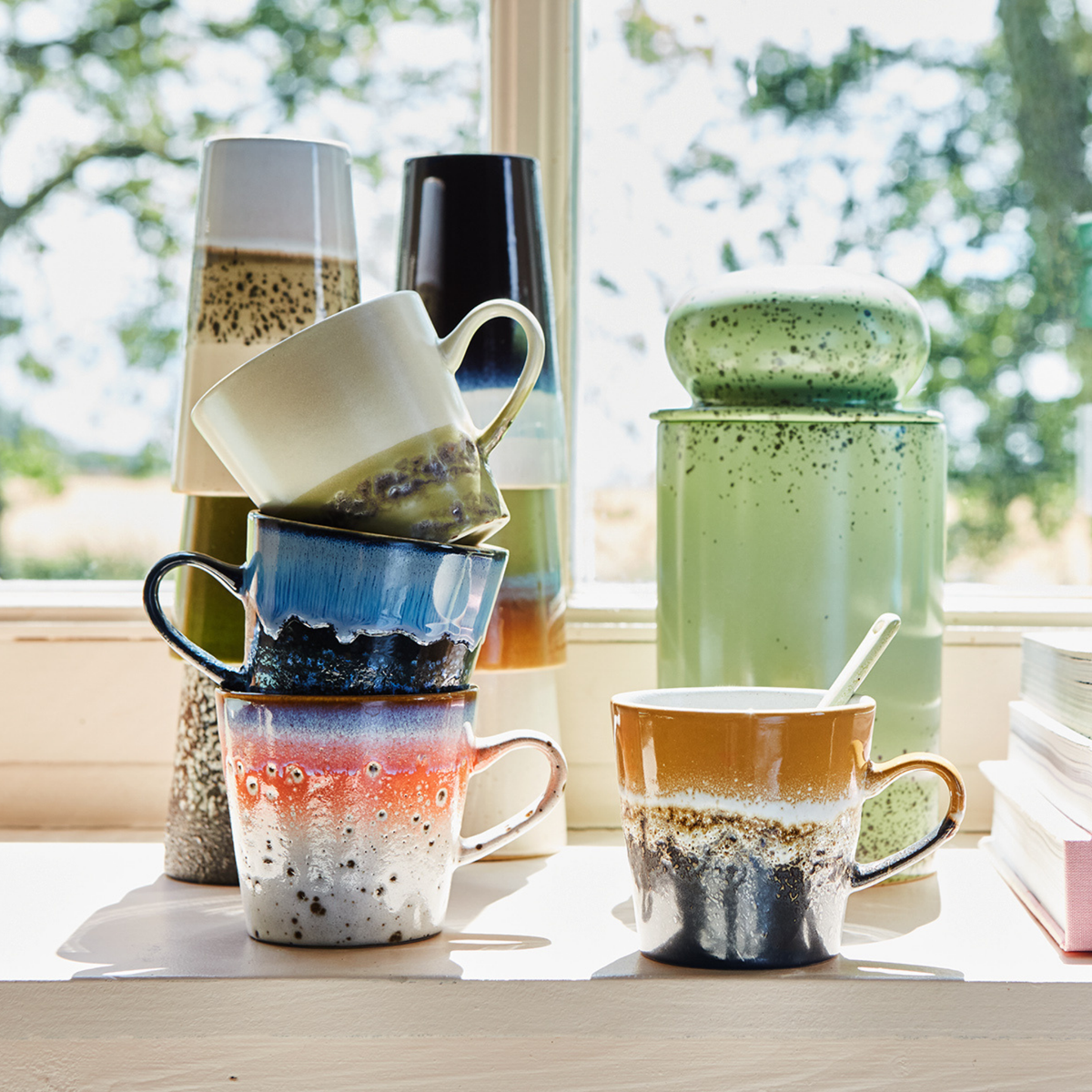 Vier HK Living 70s Ceramics Americano Mok Night-mokken staan opgestapeld op een zonnige vensterbank naast een groene glazen pot, een lepel, boeken en vazen – zo ontstaat een uitnodigend retro-koffiemoktafereel terwijl het zonlicht naar binnen stroomt.