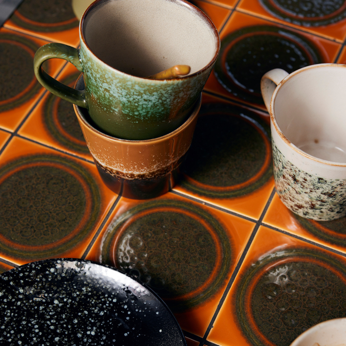 Een close-up van gestapelde HK Living 70s Ceramics Americano Mok Fire-mokken en -borden met gespikkelde, aardse patronen, uitgestald op een retro tafel met oranje en groene tegels die de klassieke HK Living-stijl weerspiegelt.