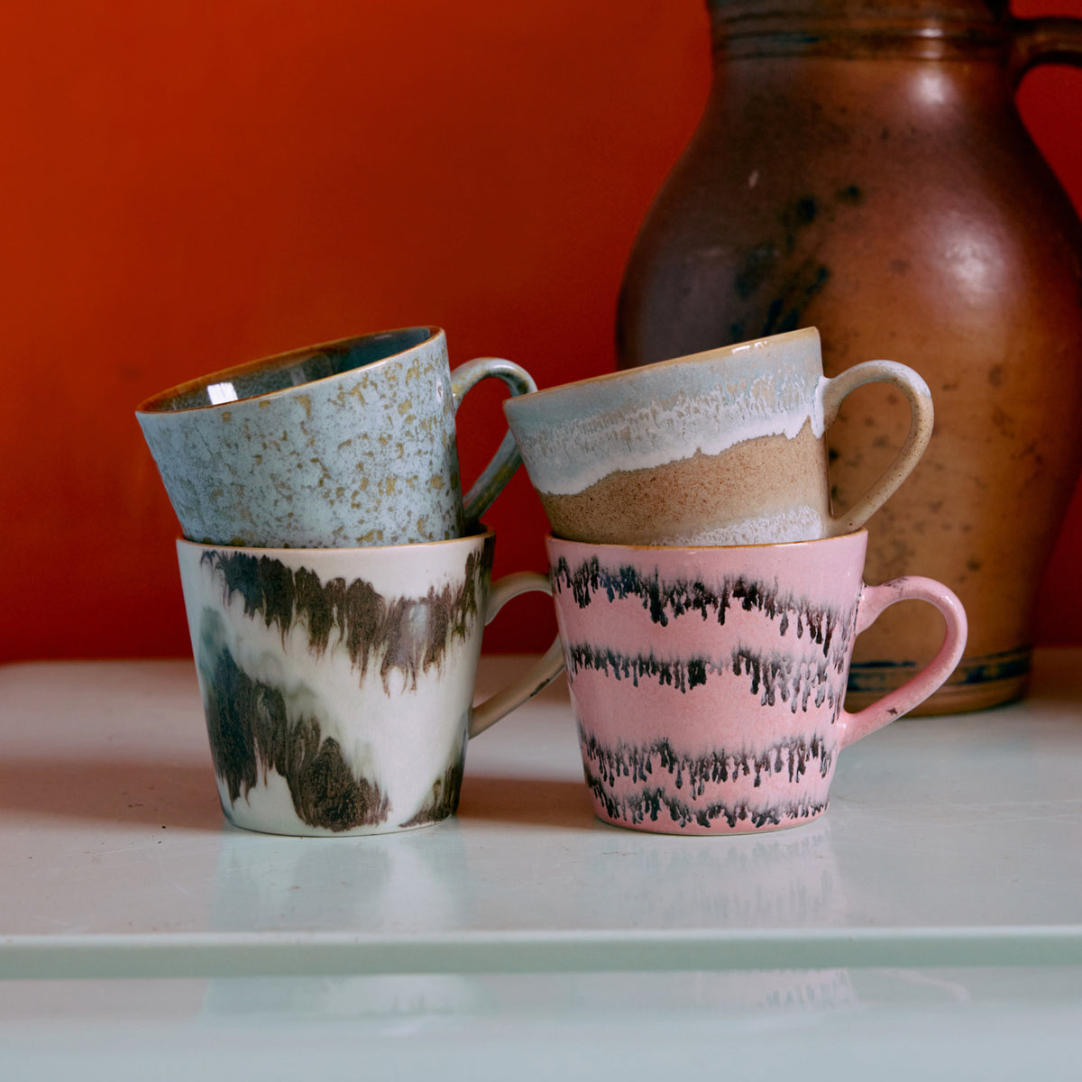 Een set HK Living 70s Ceramics Cappuccinokop Dada 4 st. mokken met unieke, kleurrijke glazuurpatronen zijn gestapeld op een wit oppervlak, met een aardebruine kan en een oranje muur op de achtergrond.