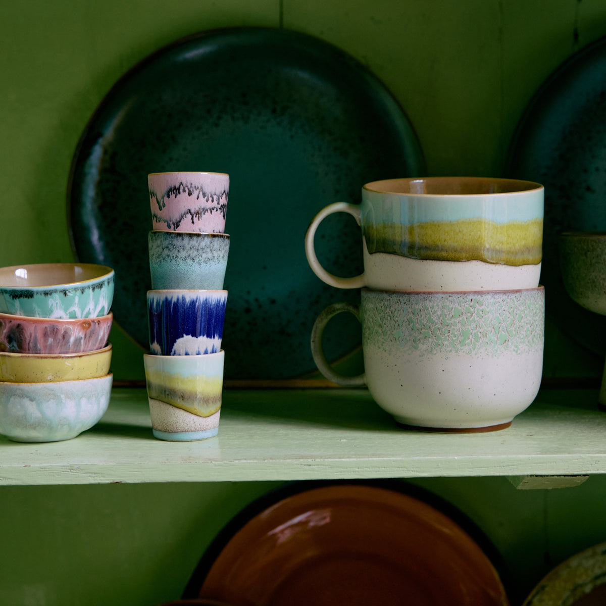 HK Living 70s Ceramics Eierdop Vibrance (set van 4) en andere kleurrijke keramische kopjes, mokken en grote borden met pastelglazuren, artistieke patronen, spikkels en retroverlopen staan op een groene plank.