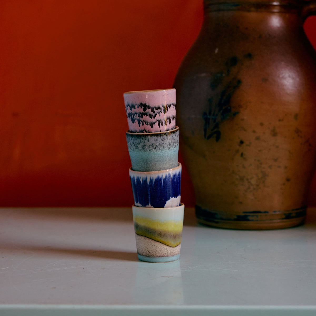 Vier HK Living 70s Ceramics Eierdop Vibrance kopjes zijn gestapeld op een bleek oppervlak voor een aardse kan en oranje wand. Elk kopje heeft een uniek, artistiek glazuur geïnspireerd op de retro eierdop stijl van HK Living.