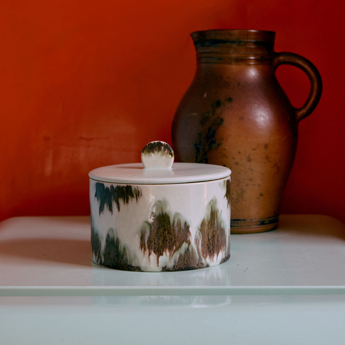 De HK Living 70s Ceramics Koekjestrommel Tide, met een bruin en groen glazuur en deksel, staat op een lichtgekleurde ondergrond voor een hoge bruine kan tegen een oranje achtergrond, en straalt de subtiele retro-charme van het merk HK Living uit.