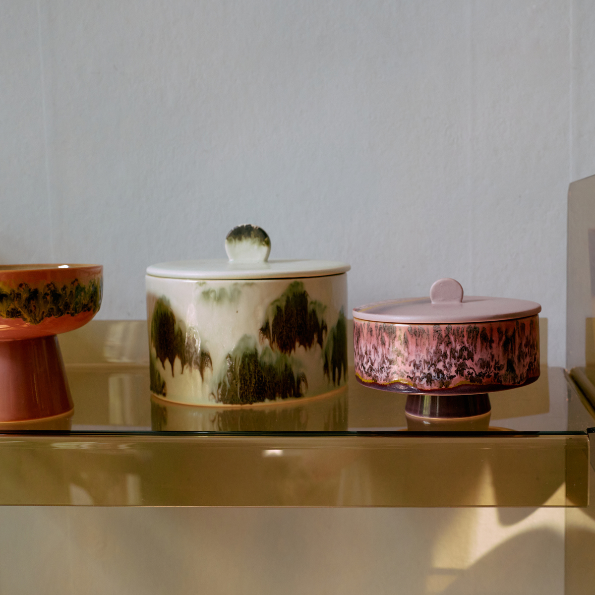 Drie HK Living 70s Ceramics Koekjestrommel Tide-bakjes met deksel, in retro gemarmerd groen, crème en roze, staan uitgestald op een glanzende gouden plank tegen een lichtgekleurde muur.