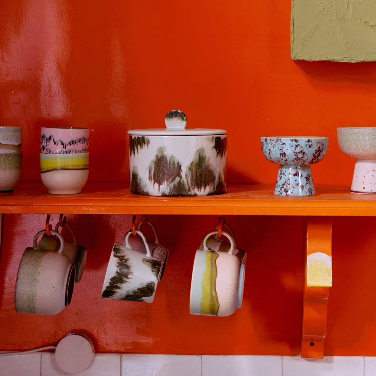 De ‘70s Ceramics Koekjestrommel Tide’ van HK Living, met groene en bruine patronen, staat op een feloranje plank tegen een oranje muur, wat zorgt voor een retro-geïnspireerde keukenlook.