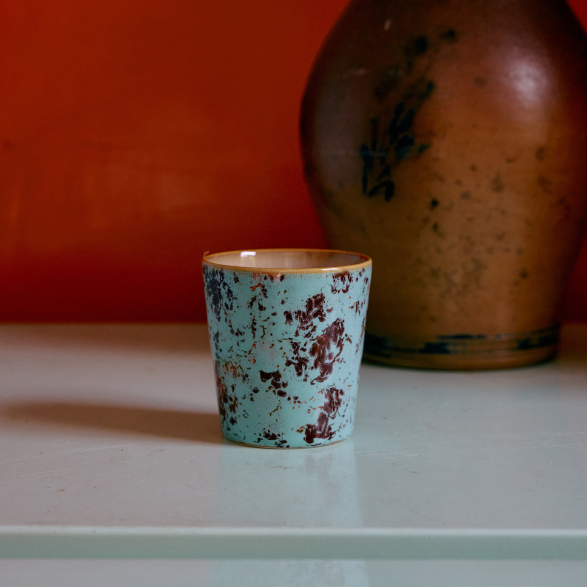 De HK Living 70s Ceramics Koffiekop Patina, een blauw met bruin gespikkelde beker van HK Living, staat op een licht oppervlak met een grote bruine vaas erachter tegen een oranje muur.