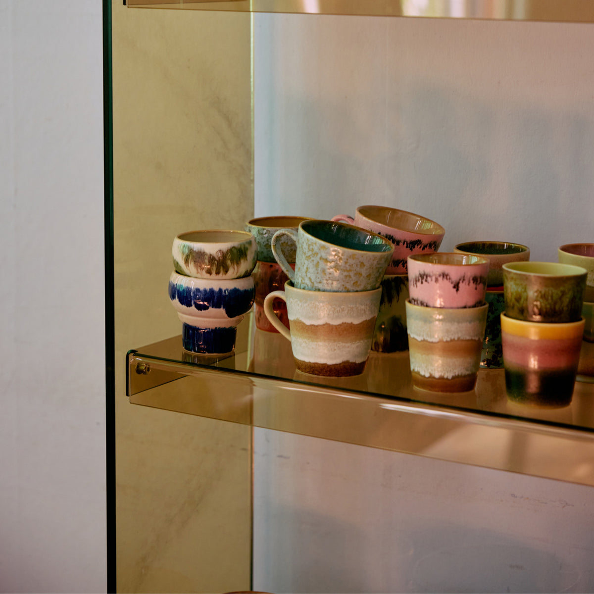 Een set levendige HK Living 70s Ceramics Lungo Mokken Expression 2 st. met unieke patronen en glazuren staat op een glazen plank tegen een goudkleurige achtergrond.