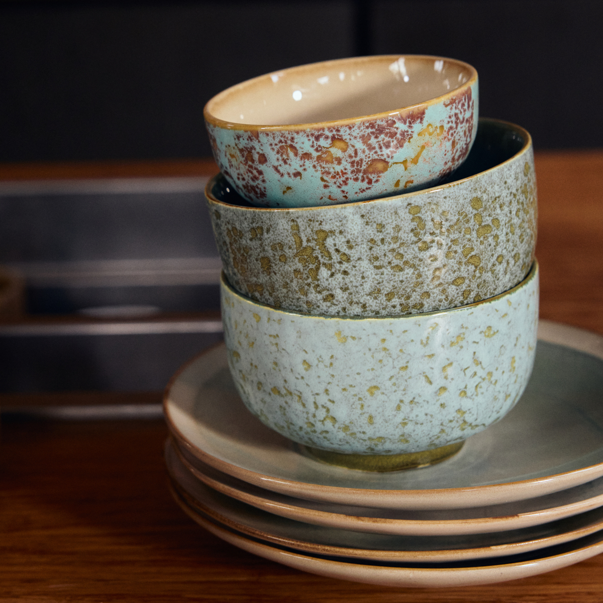 Drie HK Living 70s Ceramics Noodle Kom Verdigris D13-kommen, met blauw-groen gespikkelde patronen, staan gestapeld op bijpassende borden en geven een houten tafel een retro-uitstraling.