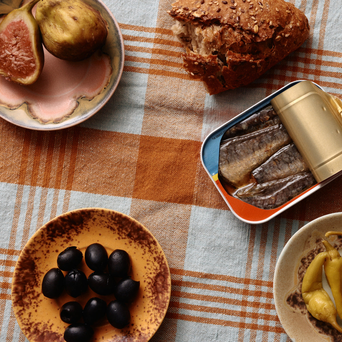 Op een tafel met een geruit oranje en wit kleed liggen sardientjes, zwarte olijven, vijgen, brood met zaadjes, ingemaakte gele paprika's en keramiek koffiekopjes naast HK Living 70s Ceramics Schotel Koffiekop Ocean D11 (set van 4).