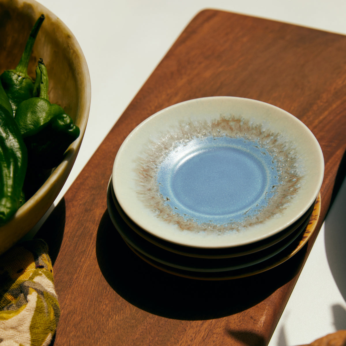 Een stapel van drie HK Living 70s Ceramics borden met blauw en bruin glazuur rust op een houten oppervlak; ernaast staat een deel van een schaal met groene paprika's. Combineer met HK Living 70s Ceramics Schotel Koffiekop Ocean D11 voor een perfecte tafelsetting.