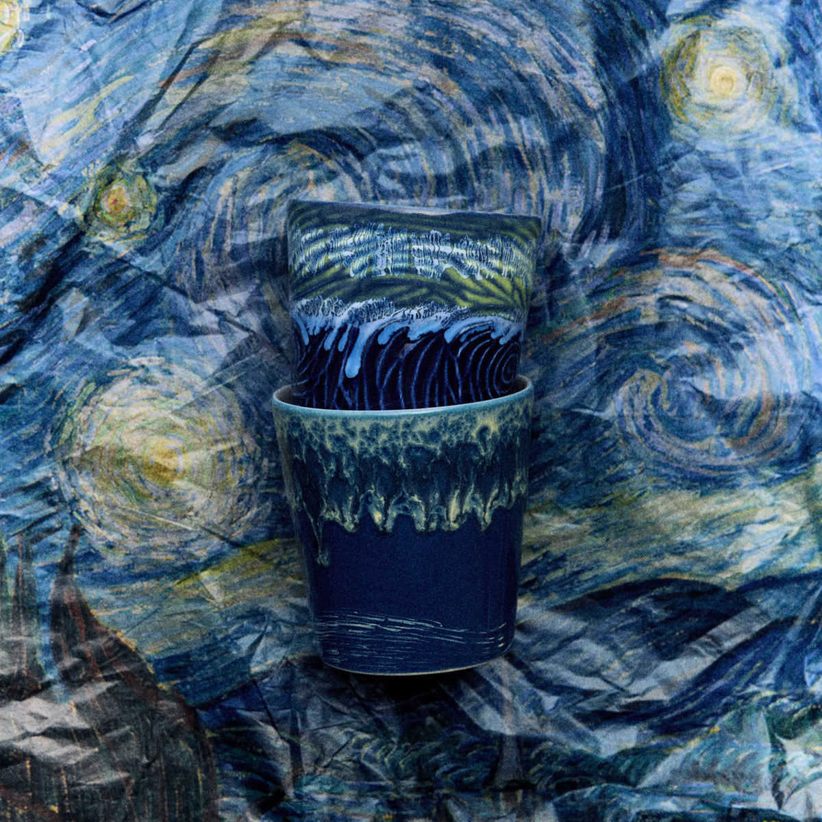 Twee blauwe HK Living 70s Ceramics Van Gogh Koffiekop Starry Night kopjes staan op elkaar gestapeld tegen een wervelende, op "Starry Night" geïnspireerde achtergrond, wat hun artistieke ontwerp en retro stijl benadrukt. Set van 2.