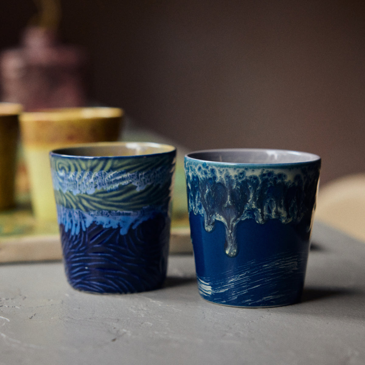 Twee blauwe HK Living 70s Ceramics Van Gogh Koffiekop Starry Night kopjes met artistieke, getextureerde glazuren rusten op een grijs oppervlak, de ene met golvend snijwerk en de andere met een glanzend, druipend ontwerp. Op de achtergrond verschijnen vage gele kopjes.