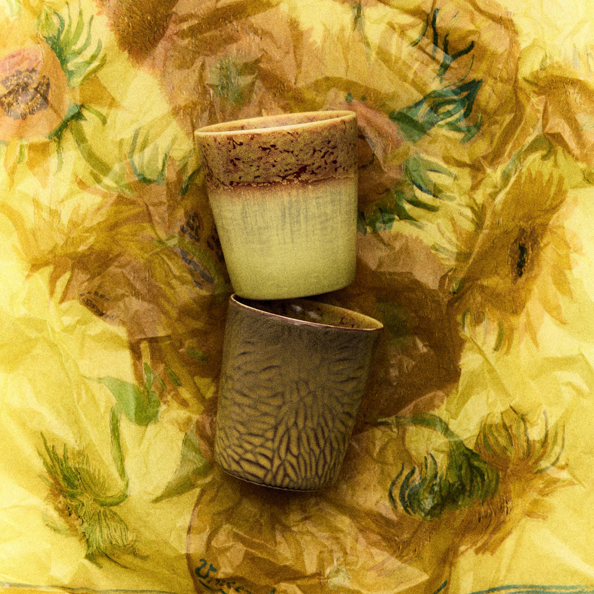 Twee HK Living 70s Ceramics Van Gogh Koffiekop Zonnebloemen kopjes met reactief glazuur zijn gestapeld op een geel oppervlak met gedurfde zonnebloemmotieven geïnspireerd op Van Goghs iconische kunst.