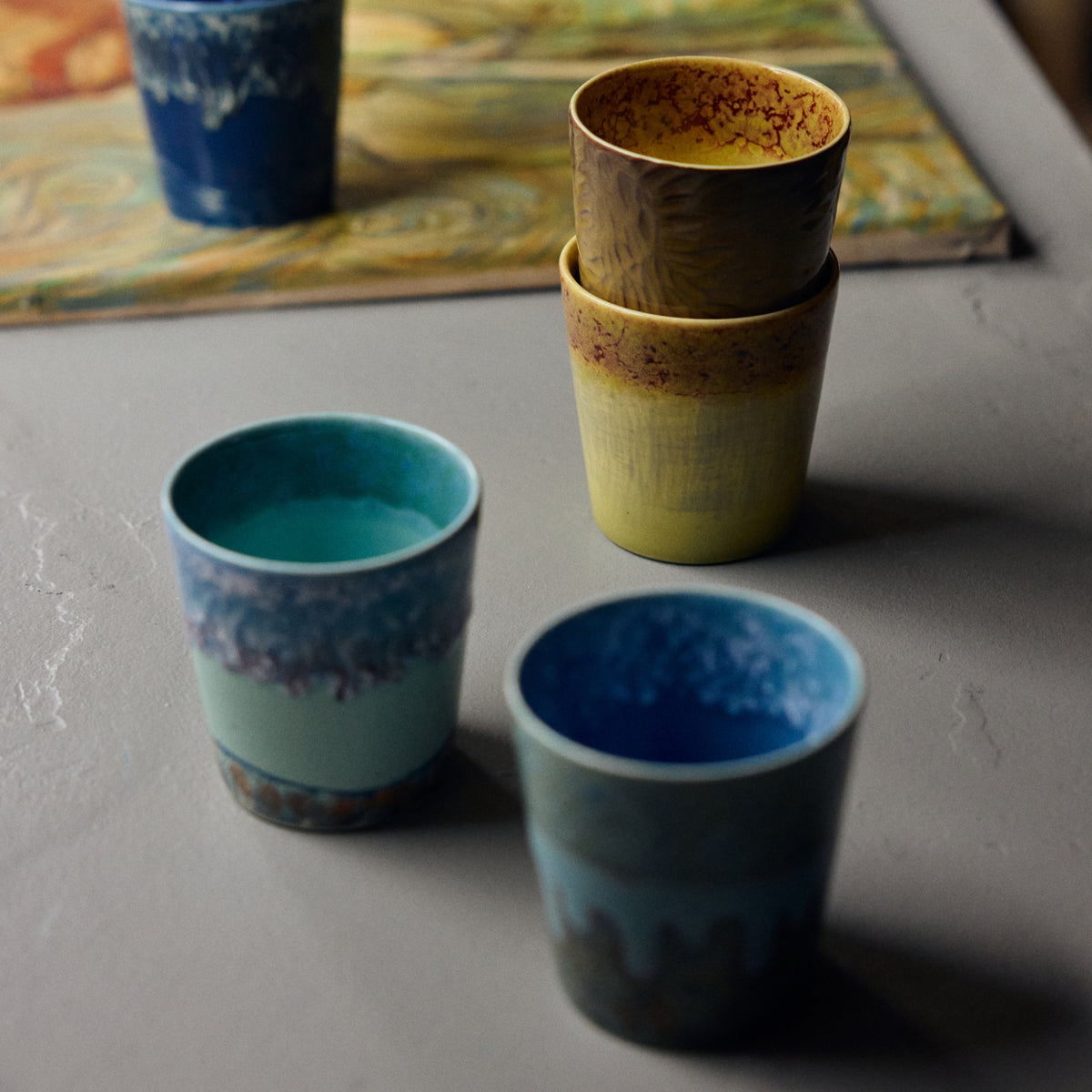 Twee HK Living 70s Ceramics Van Gogh Koffiekop Zonnebloemen kopjes met reactief glazuur staan op een grijs tafelblad; de gele en turquoise afwerkingen vallen op, met een vaag zonnebloemschilderij op de achtergrond.